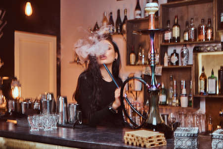 Hot brunette girl in seductive black clothes smokes a hookah while sitting on the counter in a...