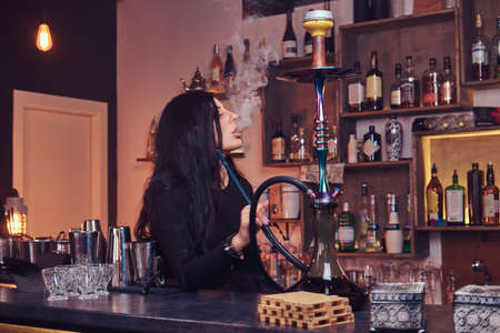Hot brunette girl in seductive black clothes smokes a hookah while sitting on the counter in a nightclub.の写真素材