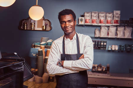 Portrait of a handsome African barista standing with crossed arms at counter of a trendy coffee shop.の写真素材