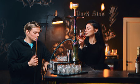 Handsome guy in hoodie smokes a hookah and seductive brunette woman drinks wine at nightclub or bar.