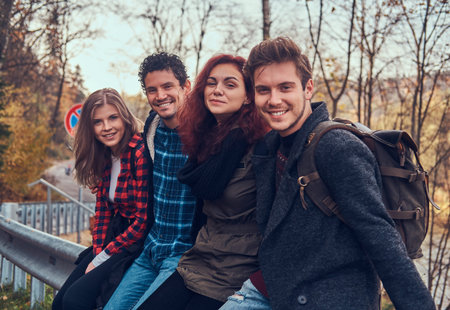Group of young friends with backpacks sitting on guardrail near road with a beautiful forest and...