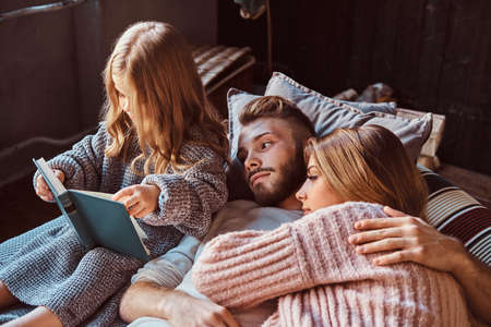 Mom, dad and daughter reading storybook together while lying on bed.の写真素材