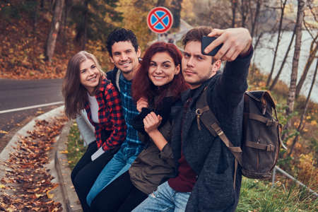 Group of young hikers with backpacks sitting on guardrail near road and doing selfie.の写真素材