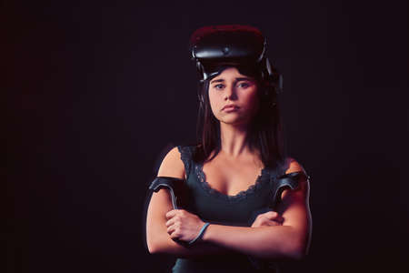 Portrait of a young woman wearing virtual reality glasses and holds joysticks, posing with crossed arms, looking at camera.の写真素材