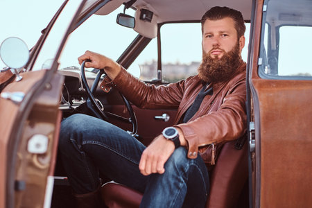 Bearded male dressed in brown leather jacket sits behind the wheel of a tuned retro car with open...