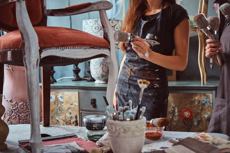 Female artist with her assistant in aprons painting vintage chair with paintbrush in workshop.