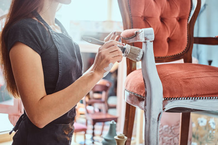 Female artist painting vintage chair in white color with paintbrush in workshop.
