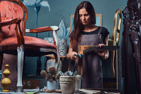 Female artist painting vintage picture frame with paintbrush in workshop.の写真素材