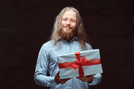 Happy male with long hair and beard in blue shirt holds gift box. Isolated on a dark background.