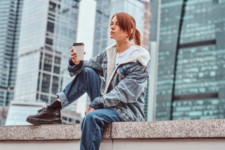 Stylish redhead hipster girl with tattoo on her face wearing denim coat holding takeaway coffee sitting in front of skyscrapers in Moskow city at cloudy morning.の写真素材