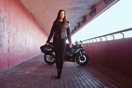 A beautiful biker girl holding helmet next superbike walks on a sidewalk inside the bridge.の写真素材