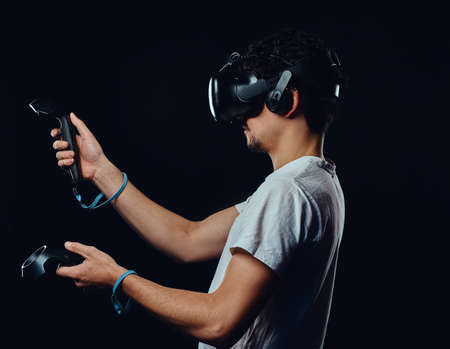 Side view of a man with bristle dressed in white shirt playing games with virtual reality goggles. Isolated on dark background.の写真素材
