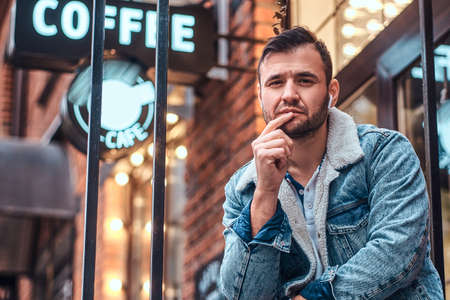 A pensive stylish man wearing a denim jacket with wireless headphones holding takeaway coffee outside the cafe.の写真素材