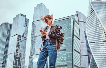 Trendy dressed redhead student girl with tattoos on her face using a smartphone in front of skyscrapers in Moskow city.の写真素材