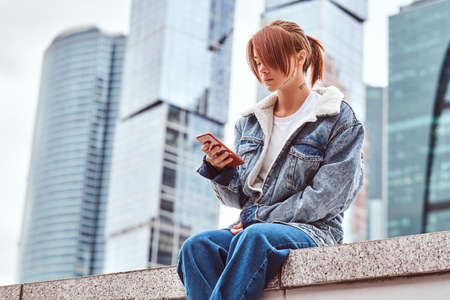 Stylish redhead hipster girl with tattoo on her face wearing denim jacket using a smartphone sitting in front of skyscrapers in Moskow city at cloudy morning.の写真素材