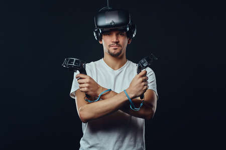 Pro gamer with bristle dressed in white shirt wearing virtual reality glasses and holds joysticks,...