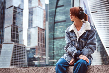 A hipster girl with tattoo on her face trendy dressed wearing headphones listening to music in front of skyscrapers in Moskow city at cloudy morning.の写真素材