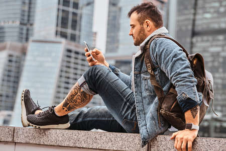 Trendy dressed tattooed hipster man with a backpack using smartphone in front of skyscrapers in Moskow city at cloudy morning.の写真素材