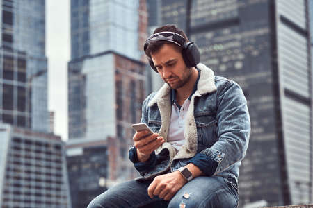 Stylish man wearing denim jacket listening to music and using smartphone sitting in front of skyscrapers in Moskow city at cloudy morning.の写真素材