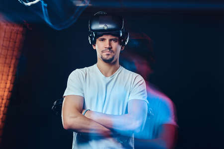 Pro gamer with bristle dressed in white shirt wearing virtual reality glasses and holds joysticks,...
