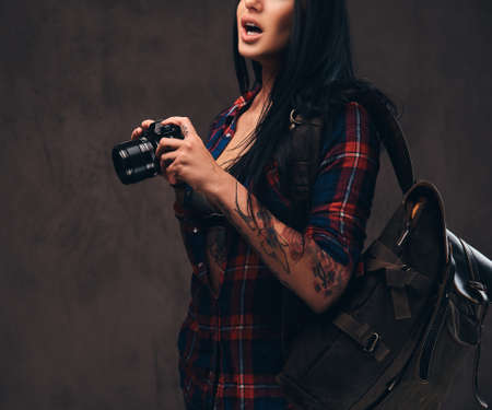 Tattooed hipster girl wearing a red unbuttoned checked shirt holding a camera on a dark background.の写真素材