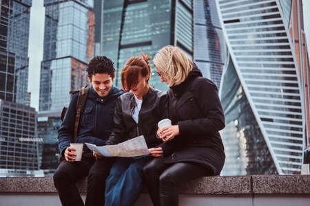 Group of tourists searching place on the map while sitting with takeaway coffee in front of skyscrapersの写真素材