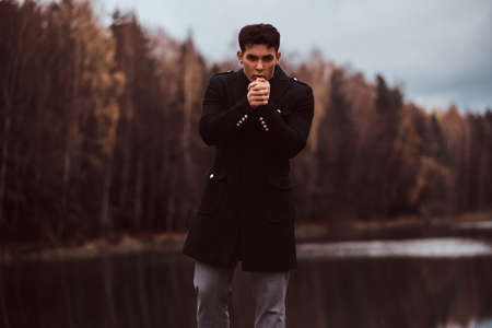 A handsome young man warms hands near a lake in the autumn forest.の写真素材