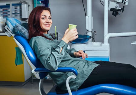 Happy woman sitting in a dentist chair holding a cup with mouthwash in a clinic.の写真素材