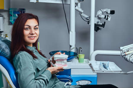 Young redhead woman sitting in a dentist chair and holding the denture in a dentist office.