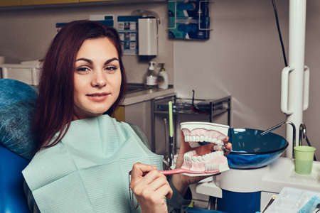 A young woman sitting in a dentist chair and holding the denture in a dentist office.