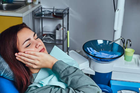 Young woman suffering from a toothache while sitting on a chair in the dentist office.の写真素材