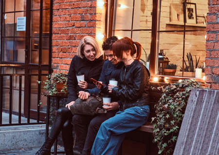 Three happy friends having a break with coffee sitting near a cafe outside.