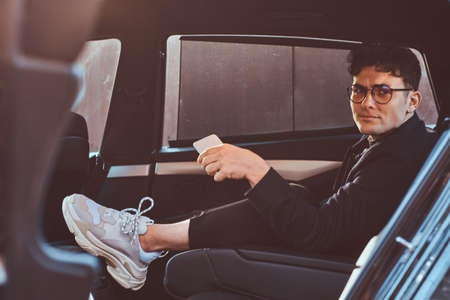 A handsome young man holding a smartphone while relaxing in the back seat of the luxury car.