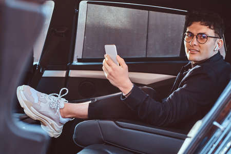A handsome young man holding a smartphone while relaxing in the back seat of the luxury car.の写真素材