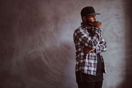 Pensive African-American bearded man standing next to a gray textured wall.