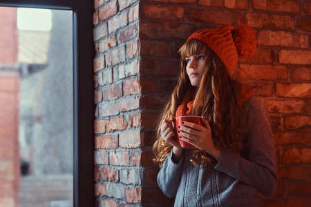 Redhead girl wearing a warm sweater and hat holding a cup of coffee while leaning on a brick wall...