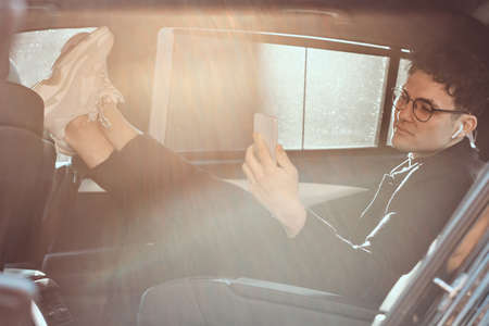 A handsome young man using a smartphone while relaxing in the back seat of the luxury car.