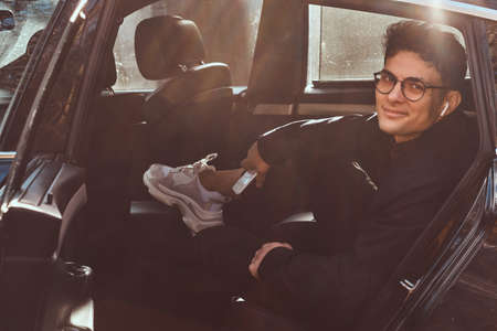 A handsome young man holding a smartphone while relaxing in the back seat of the luxury car.