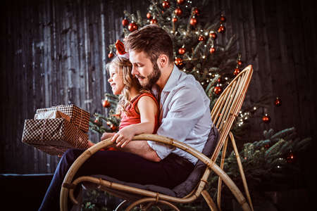Dad and daughter holding gift boxes while sitting together on a rocking chair near a Christmas tree at home.の写真素材