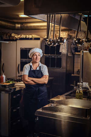 A middle age cook wearing a uniform standing with her arms crossed at restaurants kitchen.