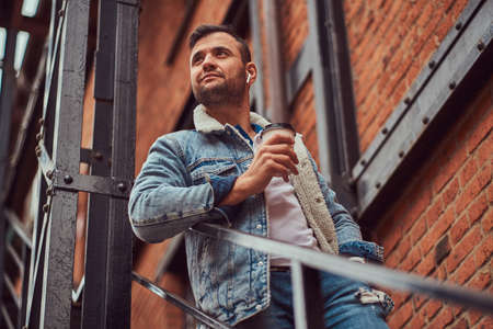 A handsome stylish man wearing a denim jacket holding a takeaway coffee on stairs outside.
