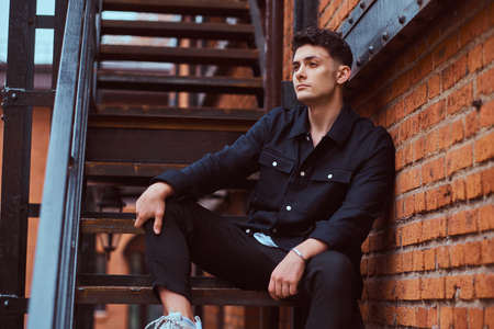 A fashionable guy sitting on the stairs outside the building with the industrial exterior.の写真素材