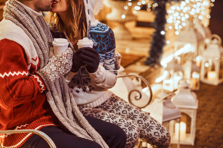 Cropped image of a young romantic couple sitting on a bench and warming with hot coffee in evening street decorated with beautiful lights at Christmas time.の写真素材