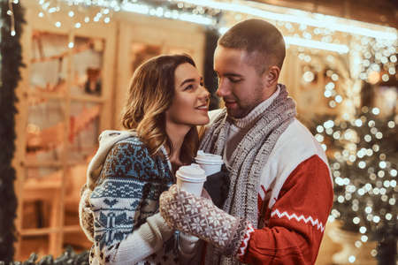A young romantic couple wearing warm clothes outdoor at Christmas time, hugging and warming with hot...