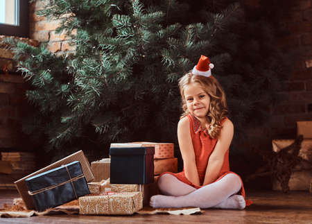 Portrait of a cute girl with blonde curly hair wearing a red dress and little Santas hat sitting on a floor surrounded by giftsの写真素材
