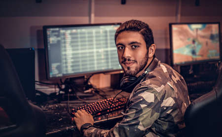 A young Indian guy wearing a military shirt sitting on a gamer chair and looking at a camera in a...