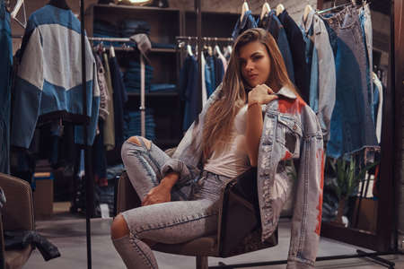 A stylish girl wearing distressed jeans and coat covering his shoulder sitting on a chair in the fitting room of a clothing store.の写真素材