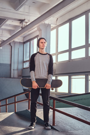 Young skateboarder standing next to a grind rail in skatepark indoors,  holding his board and looking away.の写真素材