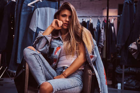A seductive girl wearing a distressed jeans and denim jacket looking at a camera while sitting on a chair in the fitting room of a clothing store.の写真素材
