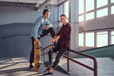 Cheerful youth couple with skateboards wearing casual clothes posing next to a grind rail in...
